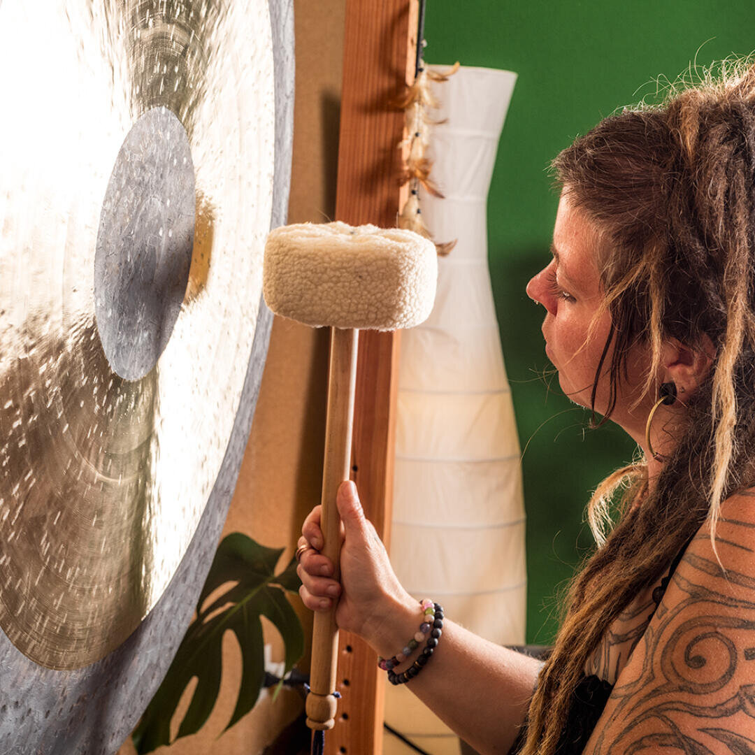 Eine Person mit langem, geflochtenem Haar hält einen gepolsterten Schlägel und bereitet sich darauf vor, einen großen Gong anzuschlagen. Im Hintergrund sind eine grüne Wand, eine weiße Lampe und eine Blattpflanze zu sehen.