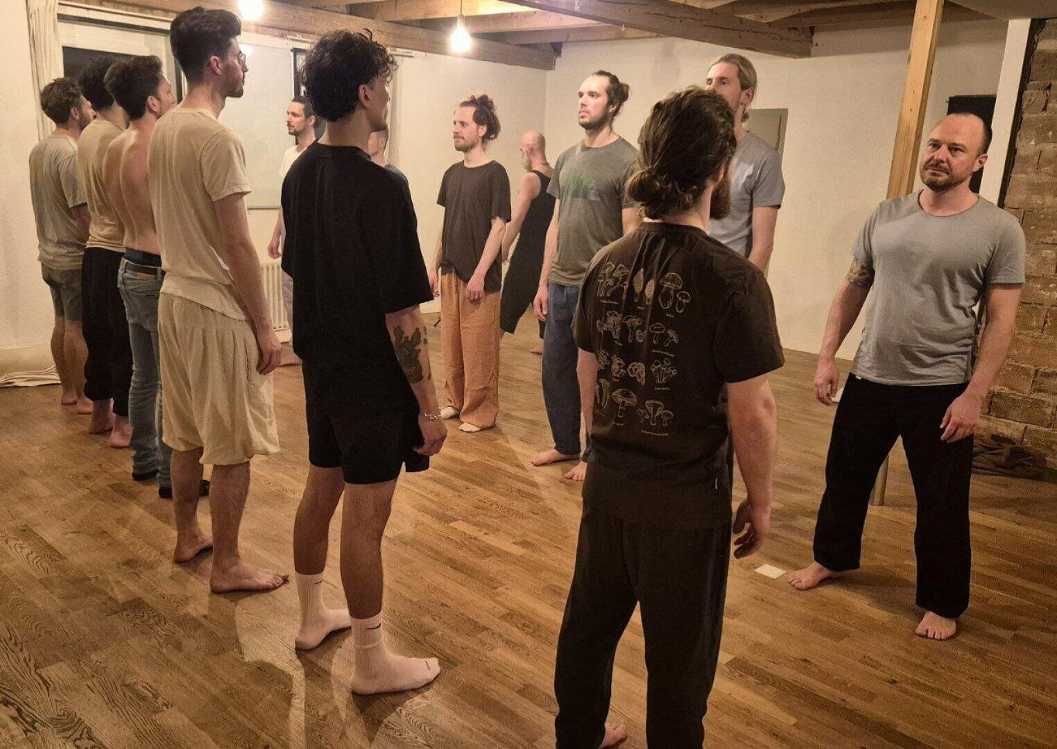 Eine Gruppe barfüßiger Männer steht sich in zwei Reihen in einem Raum mit Holzfußboden gegenüber. Sie tragen Freizeit- oder Loungekleidung und scheinen an einer Gruppenaktivität oder einem Workshop teilzunehmen.
