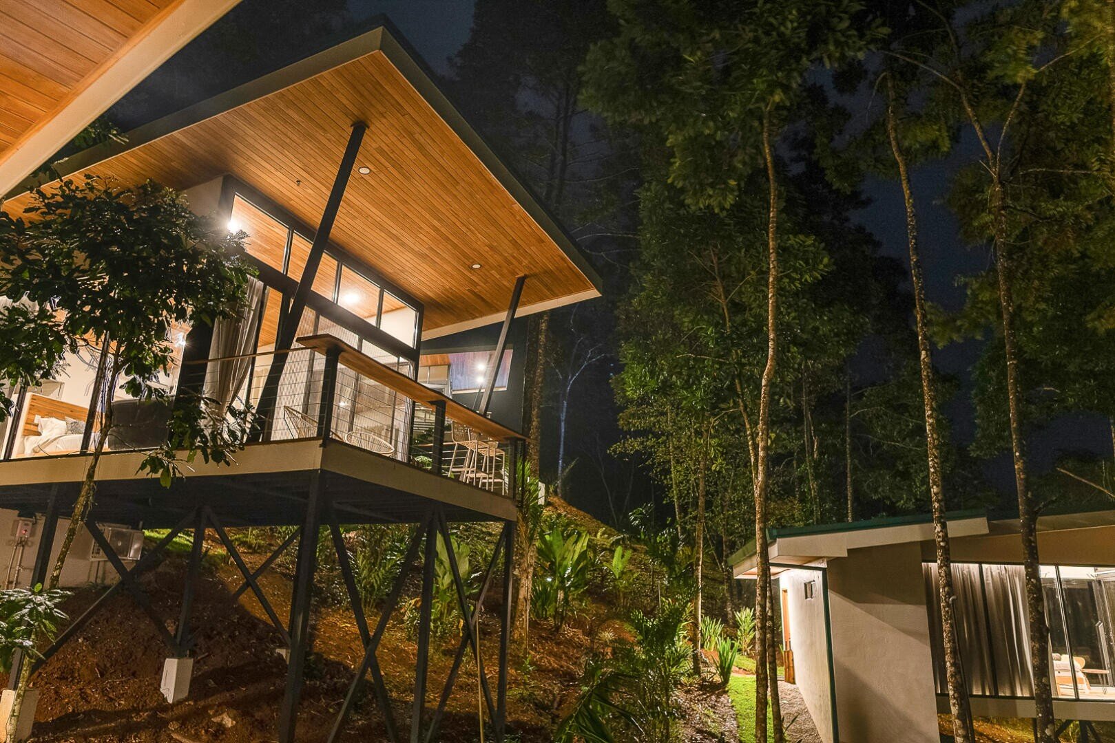 Modernes, erhöhtes Haus mit großen Fenstern und Holzdach, umgeben von hohen Bäumen bei Nacht. Warme Lichter beleuchten das Innere und das nahe gelegene einstöckige Gebäude und schaffen eine gemütliche Atmosphäre in einer Waldumgebung.