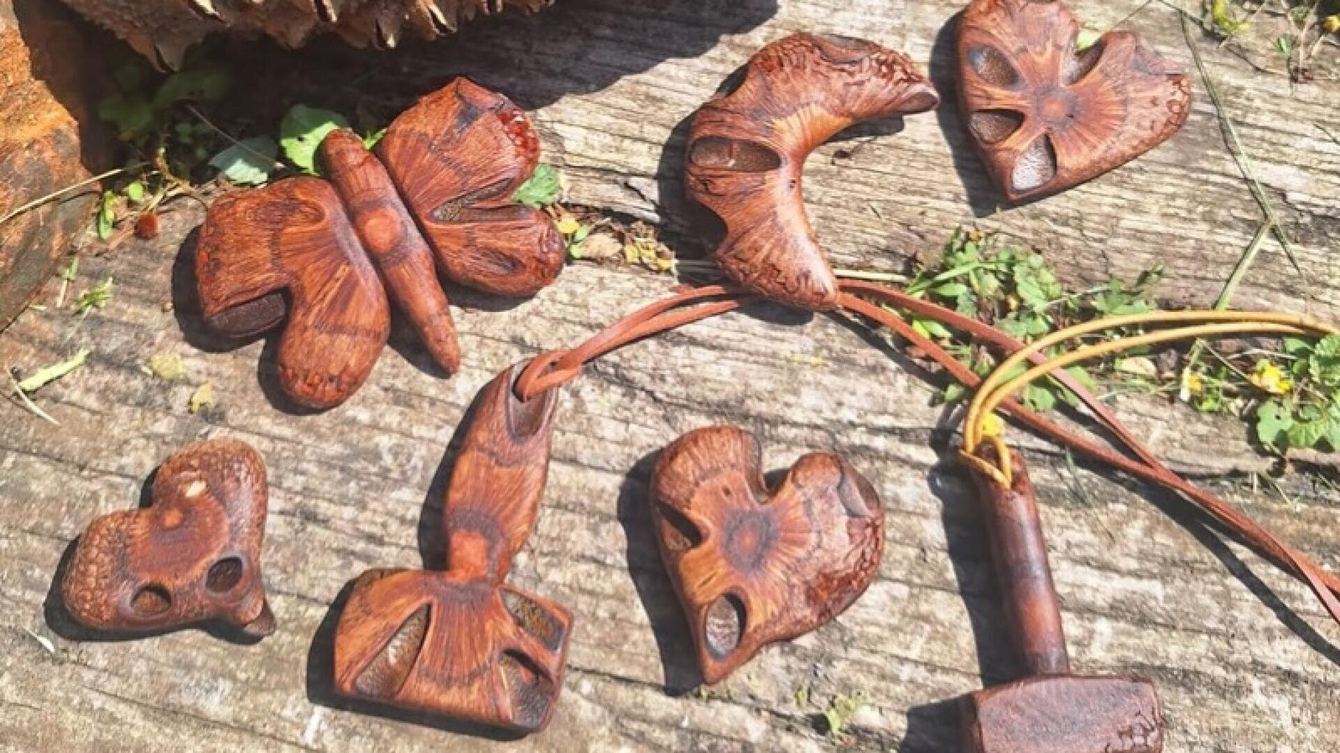 Handgeschnitzte Holzanhänger in Form von Schmetterlingen, Herzen und abstrakten Figuren sind auf einer verwitterten Holzfläche mit vereinzelten grünen Pflanzen ausgestellt.