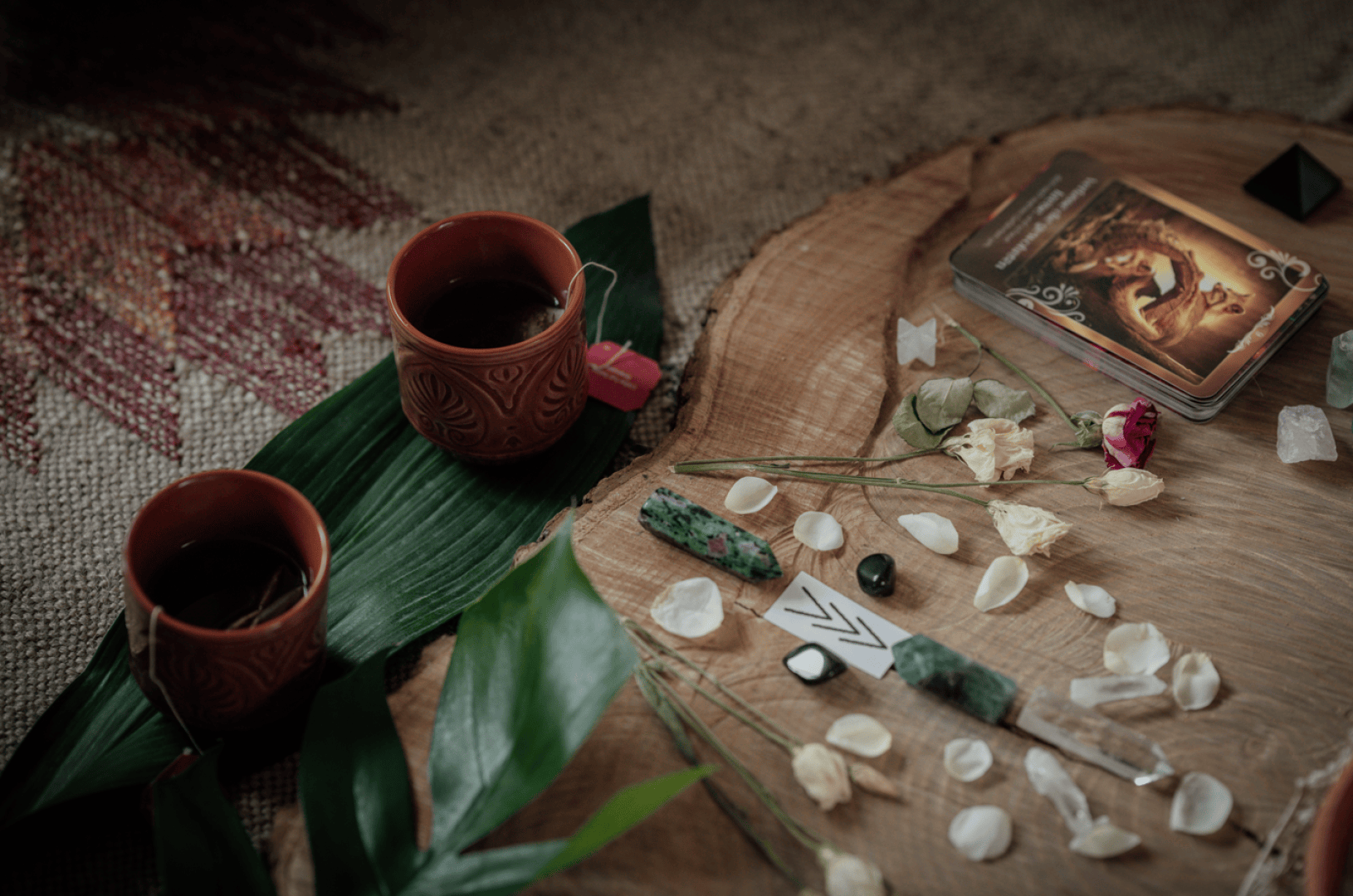 Zwei Tassen Tee, Tarotkarten, Kristalle, getrocknete Blumen und grüne Blätter sind auf einer Holzunterlage angeordnet und schaffen eine ruhige und mystische Atmosphäre.