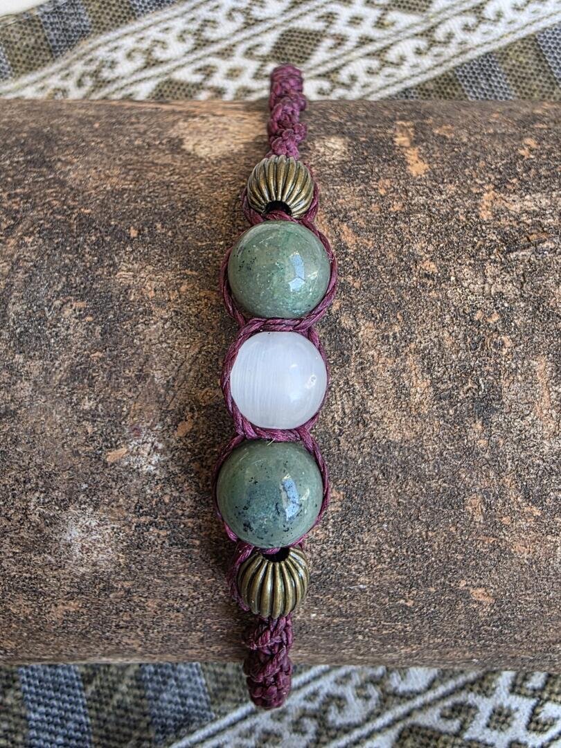 Ein geflochtenes Armband mit zwei grünen Steinen, einem weißen Stein in der Mitte und zwei runden Messingperlen, präsentiert auf einer rustikalen braunen Unterlage mit gemustertem Stoff im Hintergrund.