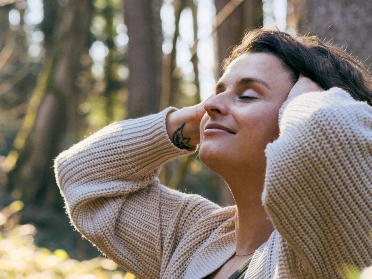 Eine Person steht in einem sonnenbeschienenen Wald, die Augen geschlossen und friedlich lächelnd, die Hände auf den Hinterkopf gestützt. Sie trägt einen leichten Strickpullover und hat ein kleines Tattoo am Handgelenk.