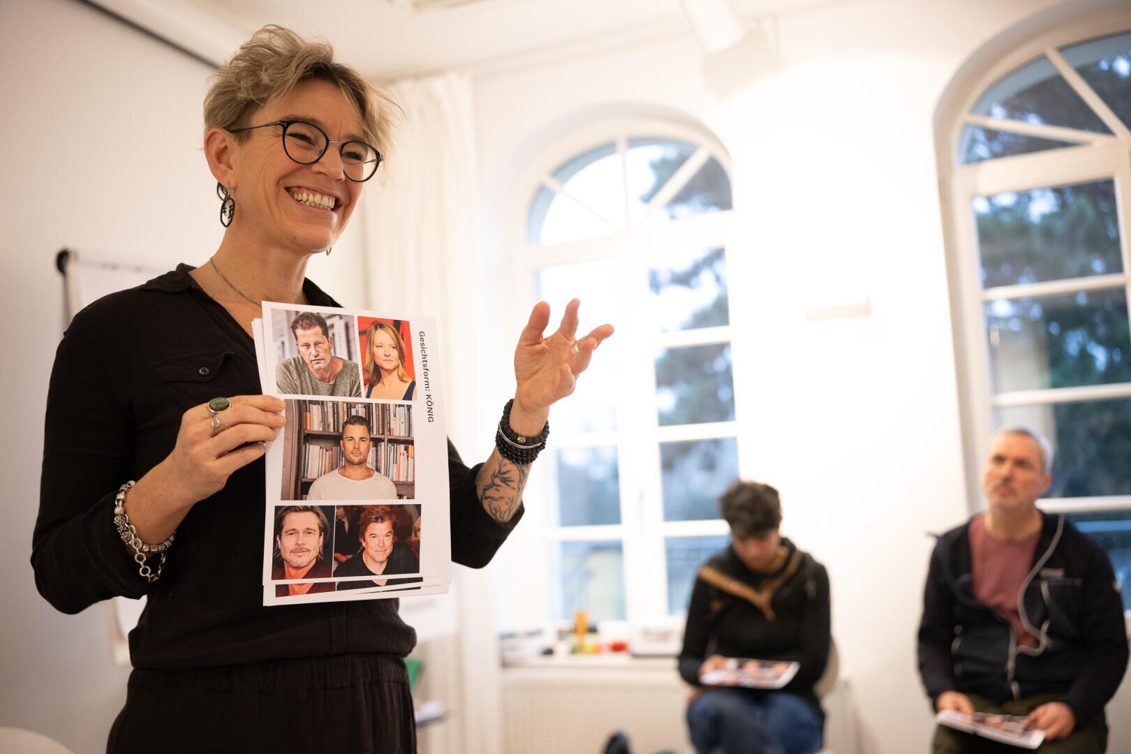In einem hellen Raum hält eine lächelnde Person mit Brille ein Blatt mit sechs Fotos von Prominenten hoch, während im Hintergrund zwei weitere Personen sitzen, von denen eine ein ähnliches Blatt in der Hand hält.