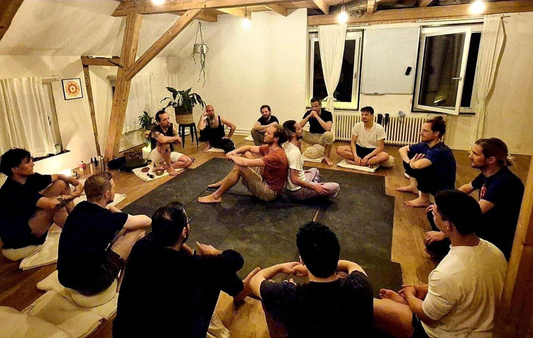 Eine Gruppe von Personen sitzt in einem Kreis auf Kissen in einem warm beleuchteten Raum und beobachtet zwei Personen in der Mitte, die anscheinend eine Partnerübung oder eine Demonstration durchführen. Die Atmosphäre wirkt ruhig und konzentriert.