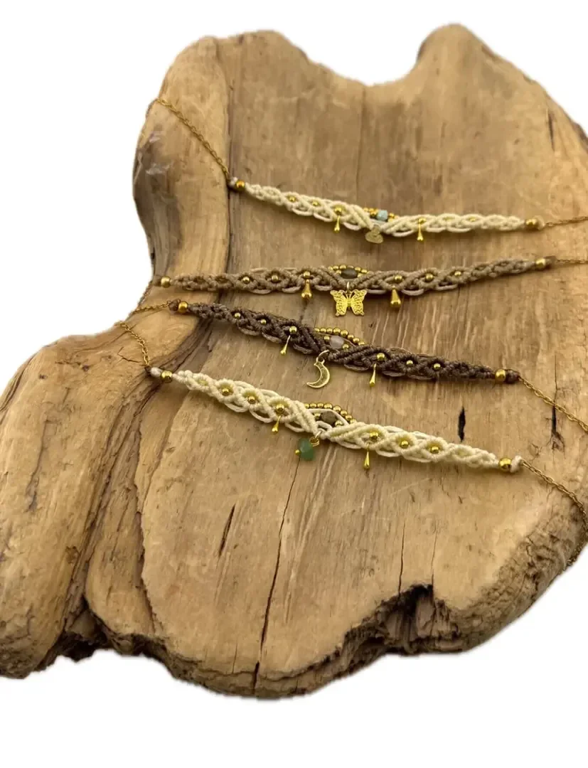 Vier handgefertigte Makramee-Armbänder mit goldenen und türkisfarbenen Perlen liegen auf einer rustikalen Holzplatte; jedes davon ist in Beige- oder Brauntönen gehalten und weist aufwendig geflochtene Muster sowie kleine Anhänger auf.