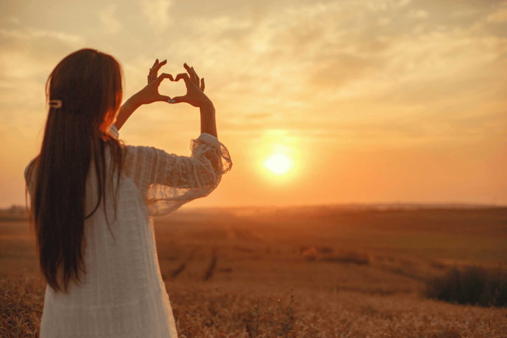 Eine Frau mit langen Haaren steht bei Sonnenuntergang auf einem Feld, der Sonne zugewandt, und formt mit ihren Händen eine Herzform. Der Himmel ist orange und golden und schafft eine warme und friedliche Atmosphäre.