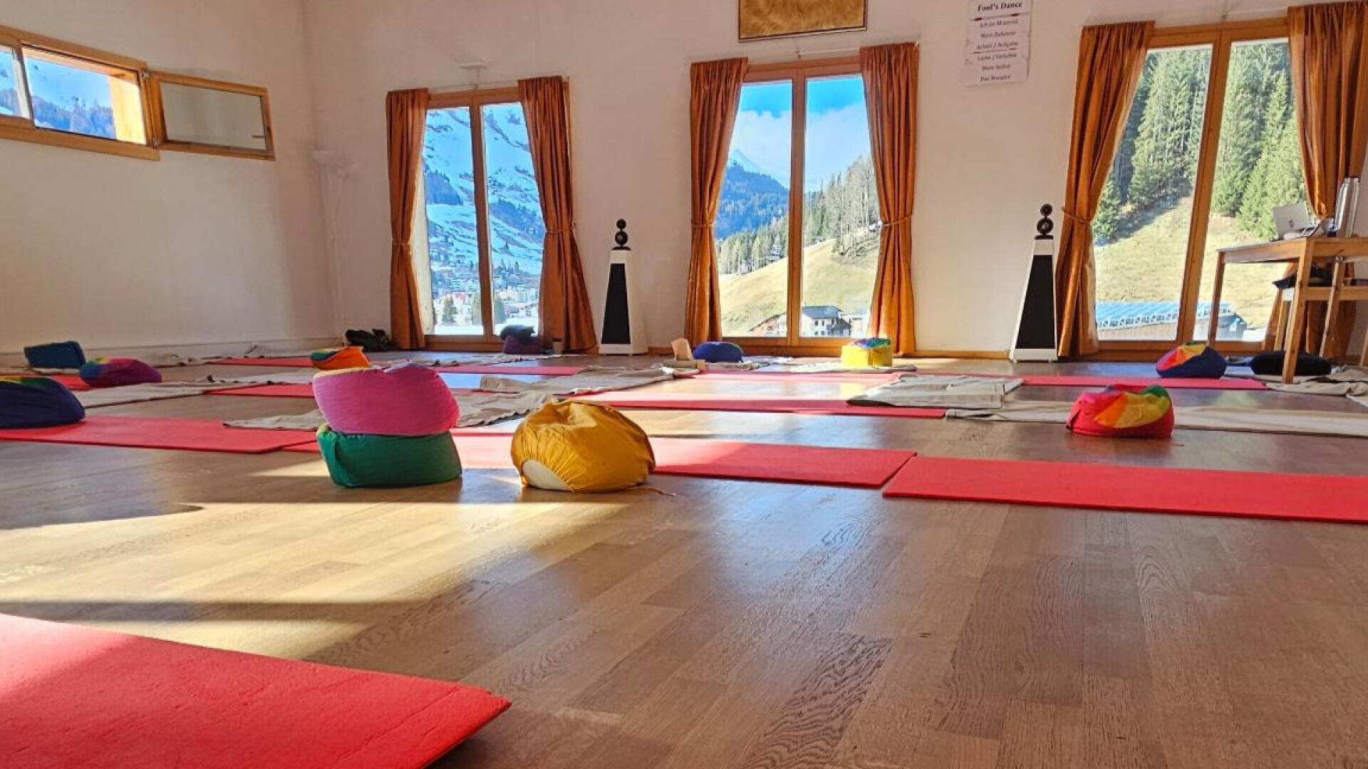 Ein sonnenbeschienenes Yogastudio mit roten Matten und bunten Kissen auf einem Holzboden. Große Fenster geben den Blick frei auf Berge, Bäume und ein kleines Dorf draußen. Der Raum ist geräumig und hell.