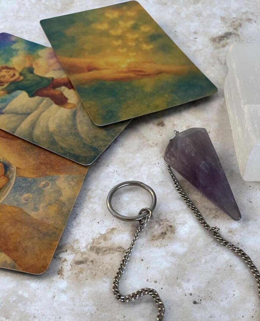 Drei illustrierte Tarotkarten, ein silberner Ring an einer Kette, ein Amethyst-Pendel und ein weißer Kristall sind auf einer hellen Marmorfläche angeordnet.