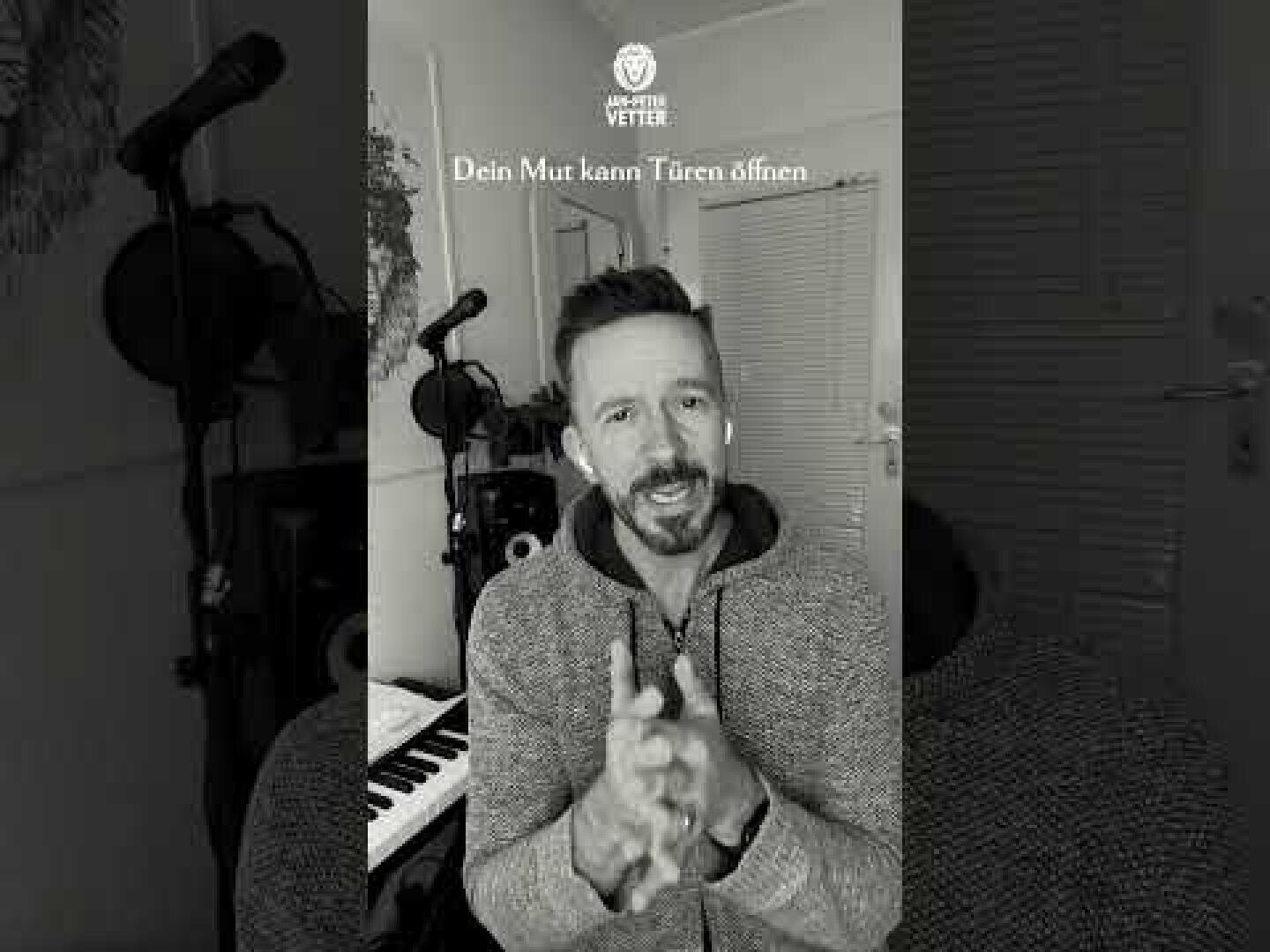 Ein Mann sitzt in einem Raum mit Musikgeräten, darunter ein Keyboard und ein Mikrofon. Er hat die Hände gefaltet, trägt Ohrstöpsel und schaut in die Kamera. Der Text über ihm lautet: Dein Mut kann Türen öffnen.