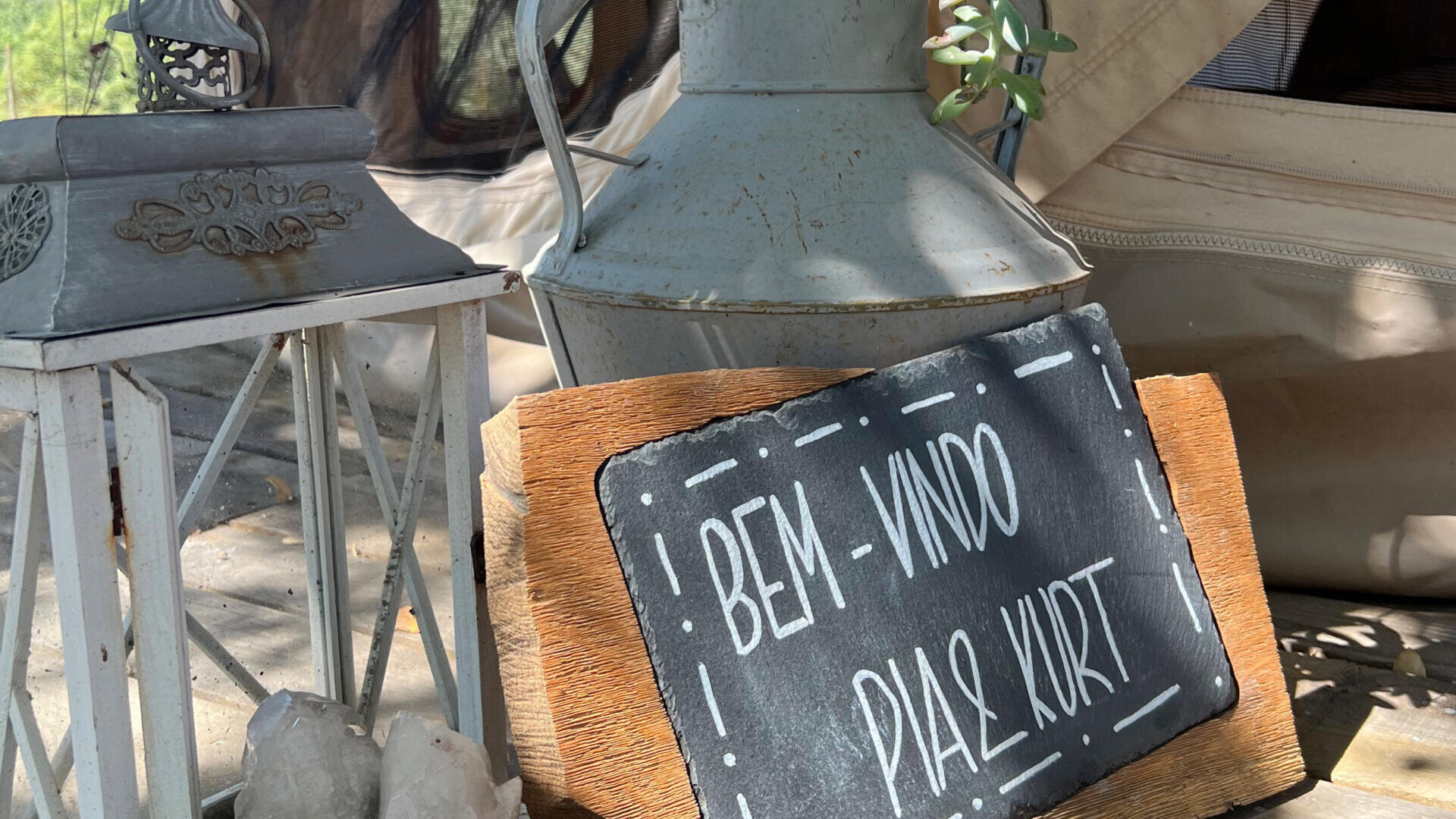 Eine Laterne, ein Metallkrug mit einer kleinen Pflanze und ein Holzschild mit der Aufschrift „BEM-VINDO PLAZA KURT“ sind draußen auf dem Boden aufgestellt, im Hintergrund ist ein Teil eines Zeltes zu sehen.