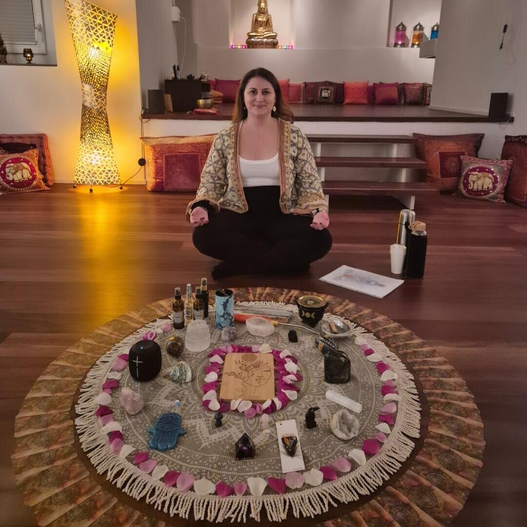 Eine Frau sitzt im Schneidersitz auf dem Boden in einer Meditationshaltung, hinter einer kreisförmigen Anordnung von Kristallen, Kerzen, Karten und spirituellen Objekten, in einem ruhigen, sanft beleuchteten Raum mit Kissen und einer Buddha-Statue im Hintergrund.