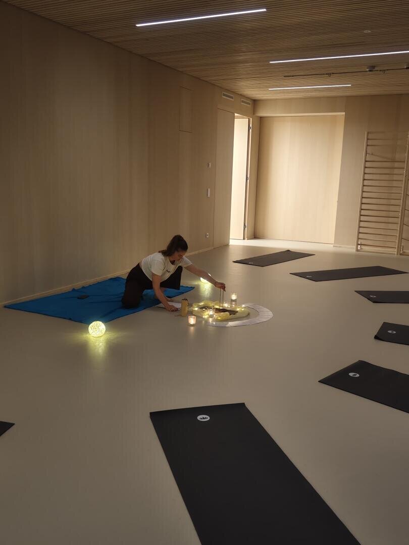 Eine Person kniet auf einer blauen Matte und arrangiert Kerzen und leuchtende Lichter in der Mitte eines geräumigen, minimalistischen Raums, der von gleichmäßig verteilten schwarzen Yogamatten umgeben ist.
