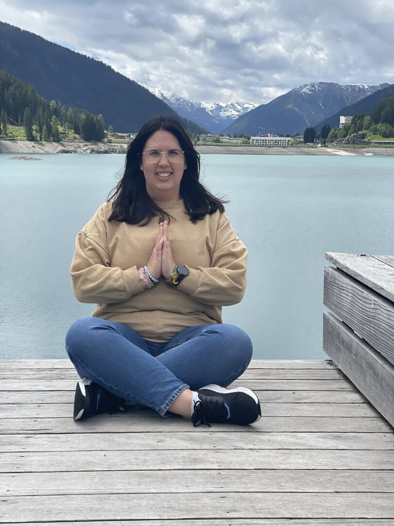 Eine Frau sitzt im Schneidersitz auf einem hölzernen Steg an einem türkisfarbenen See, die Hände in Gebetshaltung, sie trägt einen beigen Pullover und Jeans. Im Hintergrund sind verschneite Berge, grüne Hügel und ein bewölkter Himmel zu sehen.