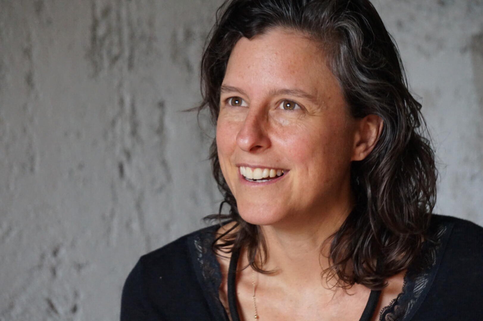 Eine Frau mit gewelltem braunem Haar und schwarzem Oberteil lächelt, während sie leicht zur Seite schaut, mit einer strukturierten grauen Wand im Hintergrund.