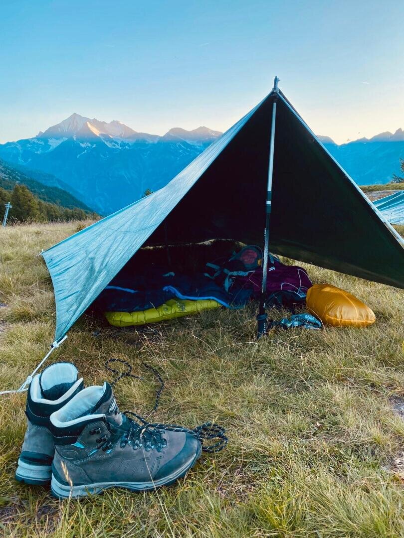 Vor einem auf grasbewachsenem Boden aufgestellten Zelt aus einer Plane stehen ein Paar Wanderstiefel; darin befinden sich ein Schlafsack und Ausrüstung. Im Hintergrund sind unter klarem Himmel Berge zu sehen.