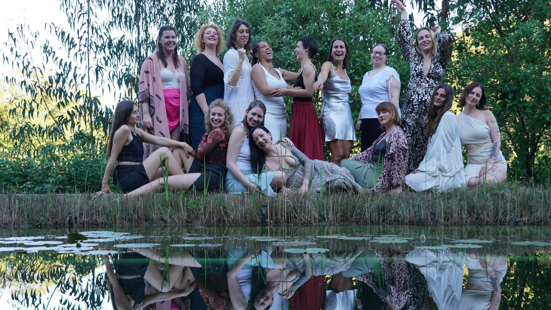 Eine Gruppe von fünfzehn Frauen posiert gemeinsam an einem von Grün umgebenen Teich, einige sitzend, andere stehend, wobei sich ihre Gesichter im Wasser spiegeln. Sie tragen eine Vielzahl von bunten und leichten Kleidern, lächeln und genießen die freie Natur.