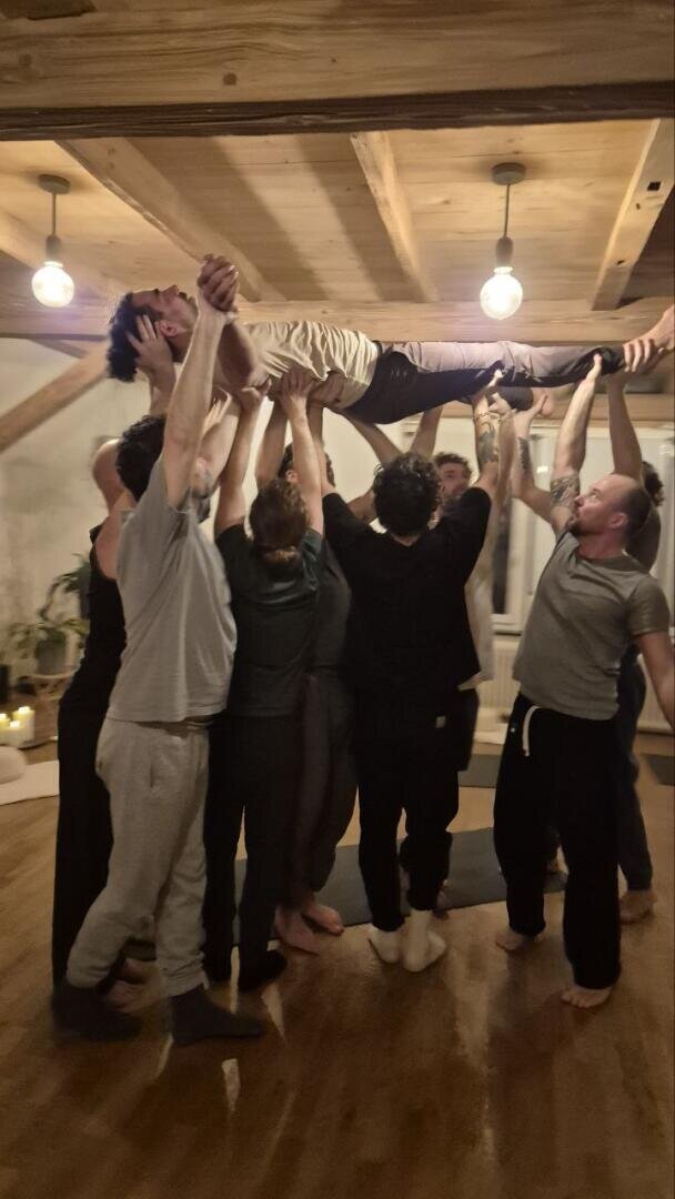 Eine Gruppe von Menschen in einem Raum hebt eine lächelnde Person über ihren Kopf und stützt sie mit ihren Armen. Der Raum hat Holzbalken, eine warme Beleuchtung und Pflanzen im Hintergrund, die eine gemütliche, gemeinschaftliche Atmosphäre schaffen.