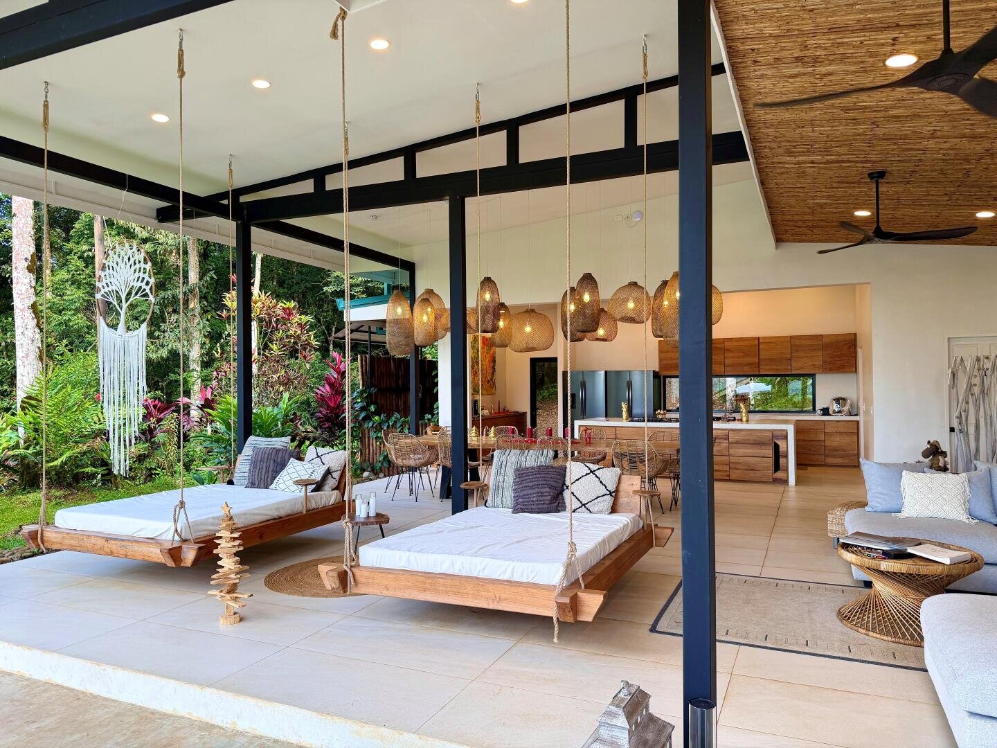 Open-Air-Wohnbereich mit Hängeliegen, modernen Holzmöbeln, geflochtenen Pendelleuchten, Zimmerpflanzen und Blick auf einen üppigen tropischen Garten vor großen Glasschiebetüren. Natürliches Licht durchflutet den stilvollen Bereich.