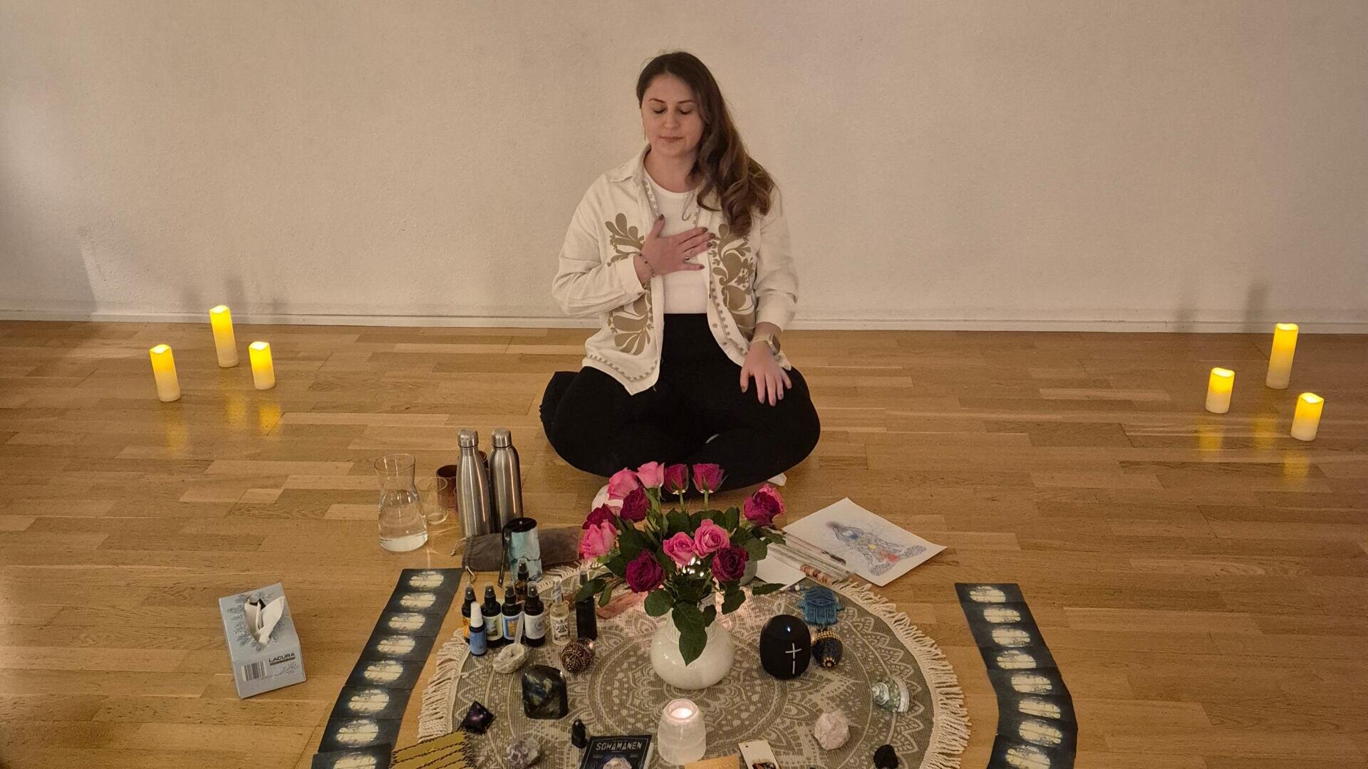 Eine Frau sitzt im Schneidersitz auf dem Boden, eine Hand auf der Brust, umgeben von brennenden Kerzen, Blumen, ätherischen Ölen und spirituellen Gegenständen, die auf einem gemusterten Tuch in einem ruhigen, sanft beleuchteten Raum angeordnet sind.