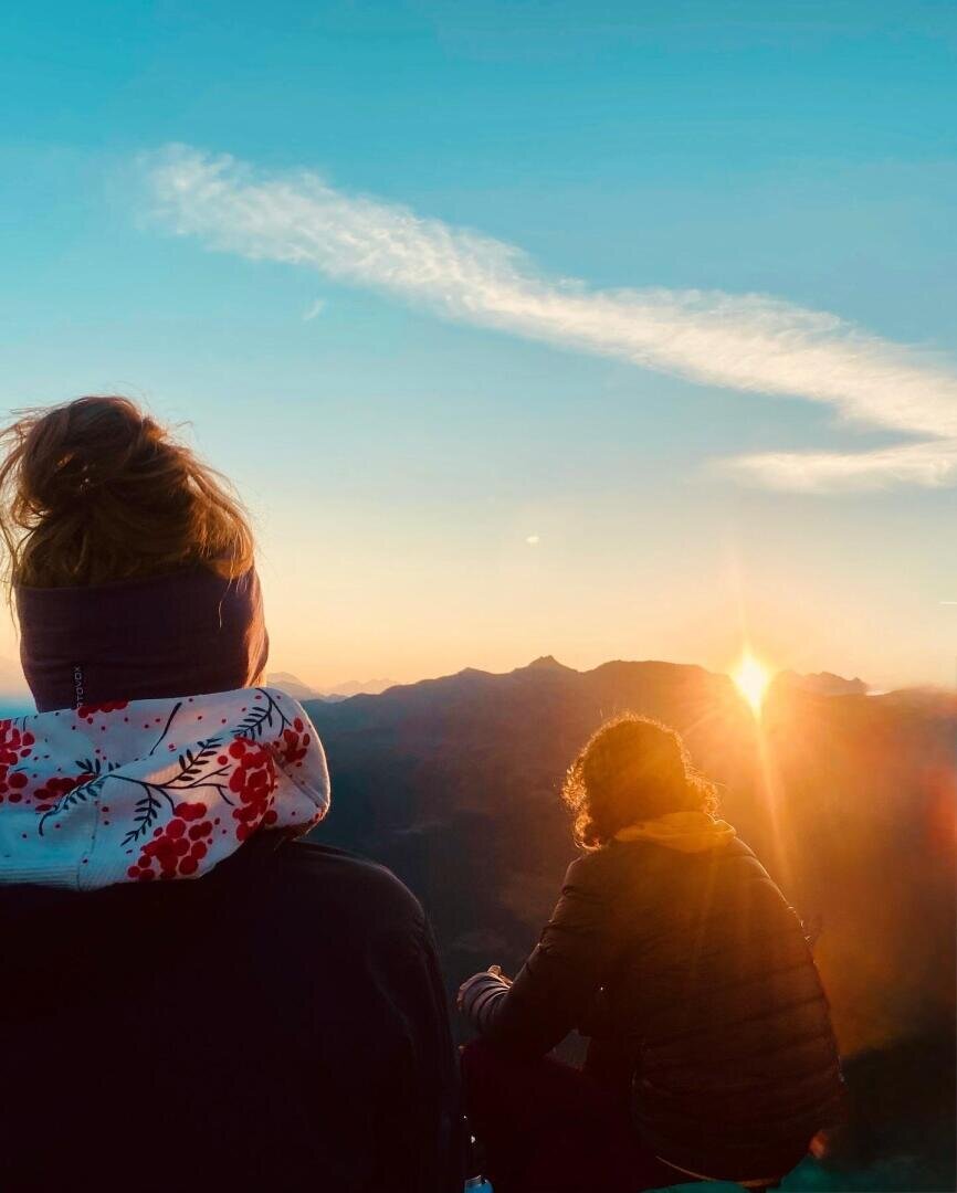 Zwei Menschen in Jacken sitzen bei Sonnenaufgang auf einem Berg und blicken in Richtung Sonne. Der Himmel ist klar, nur eine einzelne Wolke zieht vorbei, und das Sonnenlicht taucht die fernen Gipfel in einen warmen Schein.