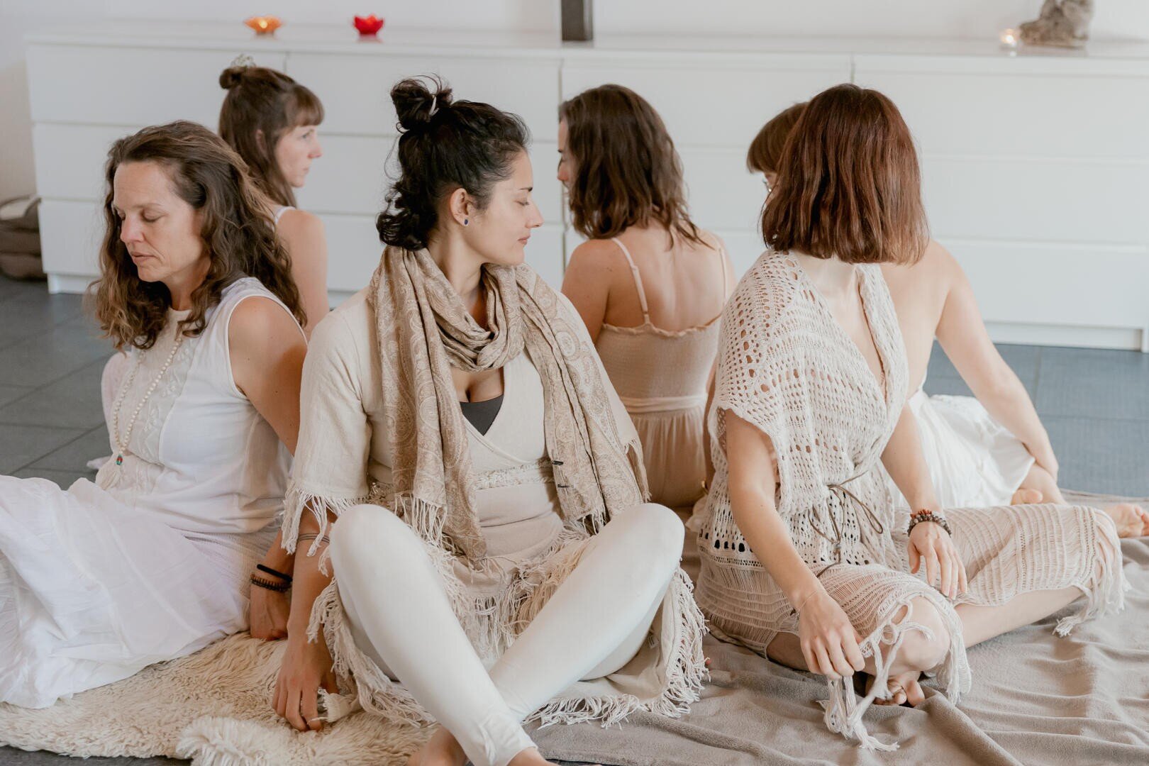 Sechs Frauen in heller, neutraler Kleidung sitzen Rücken an Rücken im Kreis auf dem Boden, die Augen geschlossen oder abgewandt, in einem ruhigen, sanft beleuchteten Raum, was an Meditation oder eine Achtsamkeitsgruppe denken lässt.