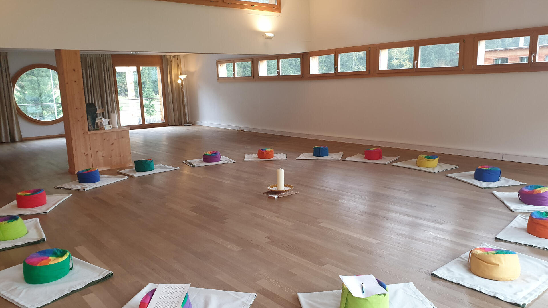 Ein geräumiger, heller Raum mit Holzböden und großen Fenstern. Bunte runde Kissen und Matten sind in einem Kreis um eine Kerze in der Mitte angeordnet, was auf eine Meditation oder ein Gruppentreffen hindeutet.