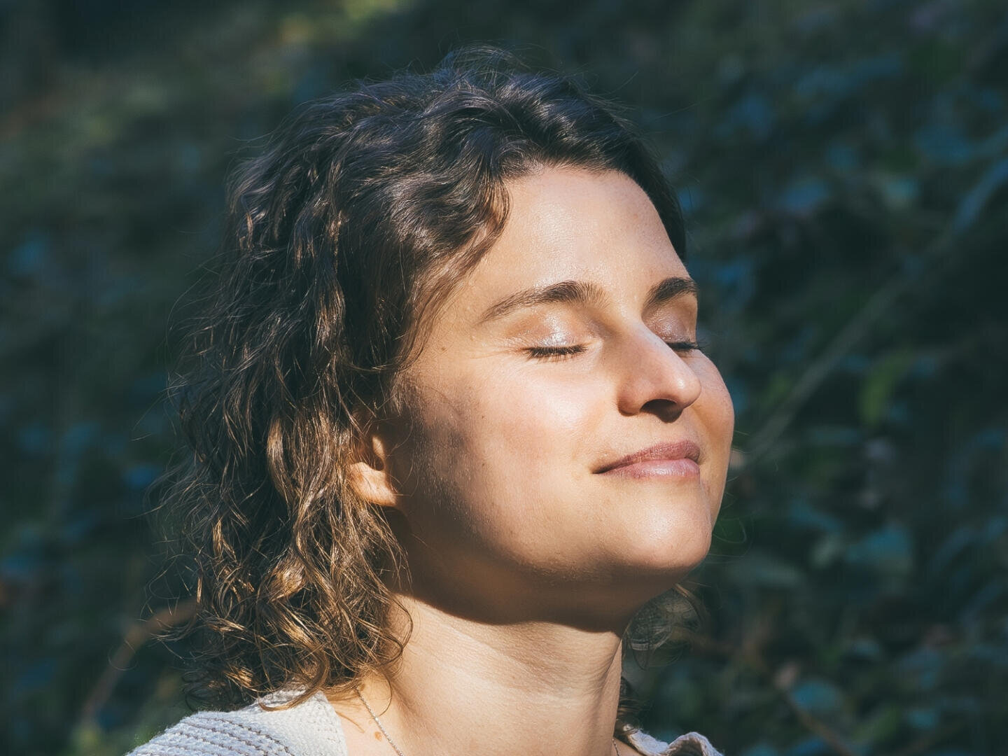 Eine Frau mit lockigem, braunem Haar lächelt sanft mit geschlossenen Augen, während sie sich im Freien im Sonnenlicht sonnt. Das grüne Laub ist im Hintergrund verschwommen.
