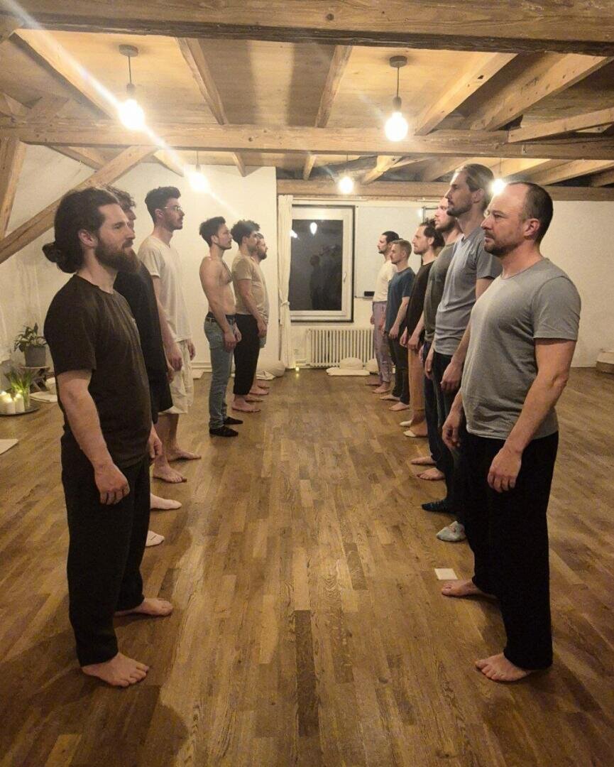 Eine Gruppe von Männern steht barfuß in zwei gegenüberliegenden Reihen auf einem Holzboden in einem Raum mit freiliegenden Holzbalken und Hängelampen und bildet eine symmetrische Formation.