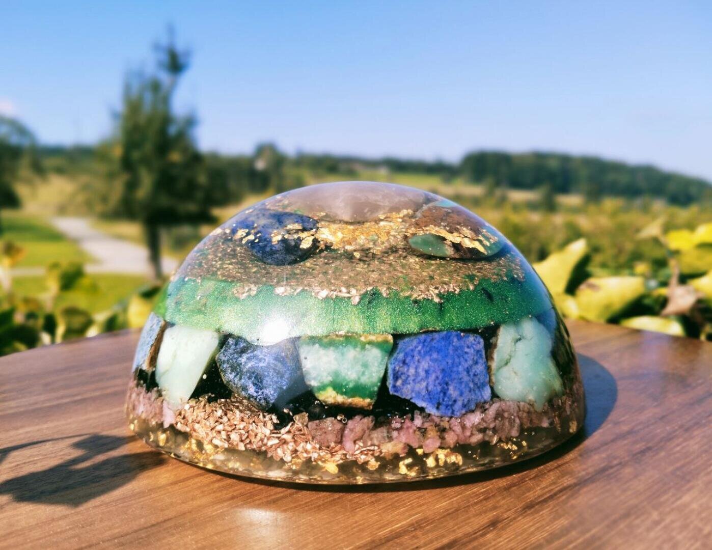 Ein kuppelförmiges Orgonit mit Schichten aus blauen, grünen und violetten Steinen sowie Goldflocken, das im Freien auf einer Holzunterlage platziert ist, im Hintergrund sind Grünpflanzen und der blaue Himmel zu sehen.
