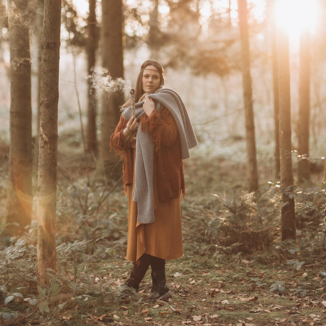Eine Frau in einem langen braunen Kleid und Stiefeln steht in einem sonnenbeschienenen Wald, hält einen grauen Schal in der Hand und atmet in der kühlen Luft sichtbaren Atem aus.