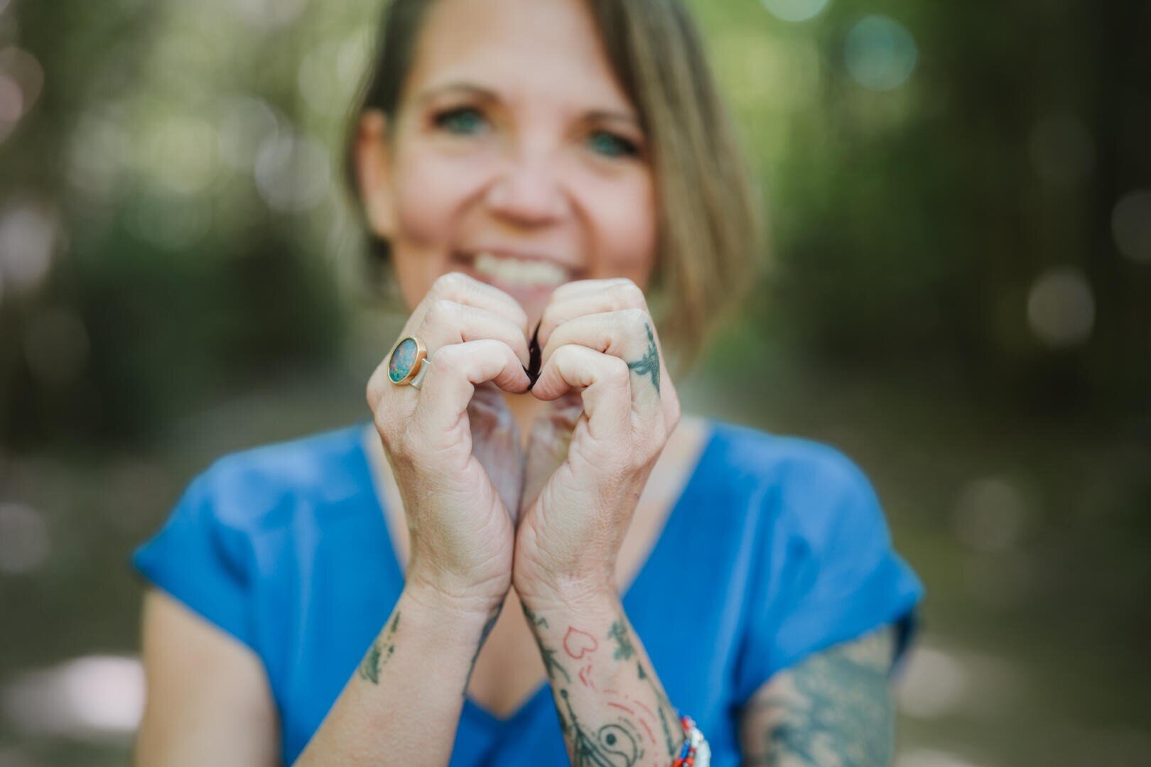 Eine lächelnde Person mit Tätowierungen und blauem Hemd formt mit ihren Händen, die sie dicht vor ihr Gesicht hält, eine Herzform in einer unscharfen Naturkulisse.