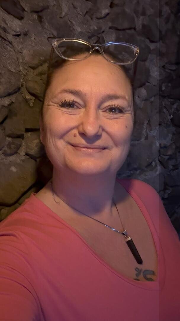 Eine Frau, die ein rosa Oberteil trägt und eine Brille auf dem Kopf hat, lächelt in die Kamera.