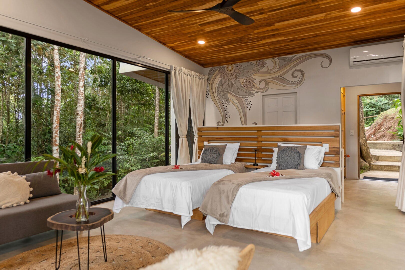 Ein modernes Schlafzimmer mit zwei Einzelbetten, neutraler Bettwäsche, dekorativen Kissen und einer Holzdecke. Große Fenster geben den Blick auf den Wald frei. Es gibt einen runden Tisch mit Blumen und ein Sofa auf der linken Seite sowie ein Wandbild über den Betten.