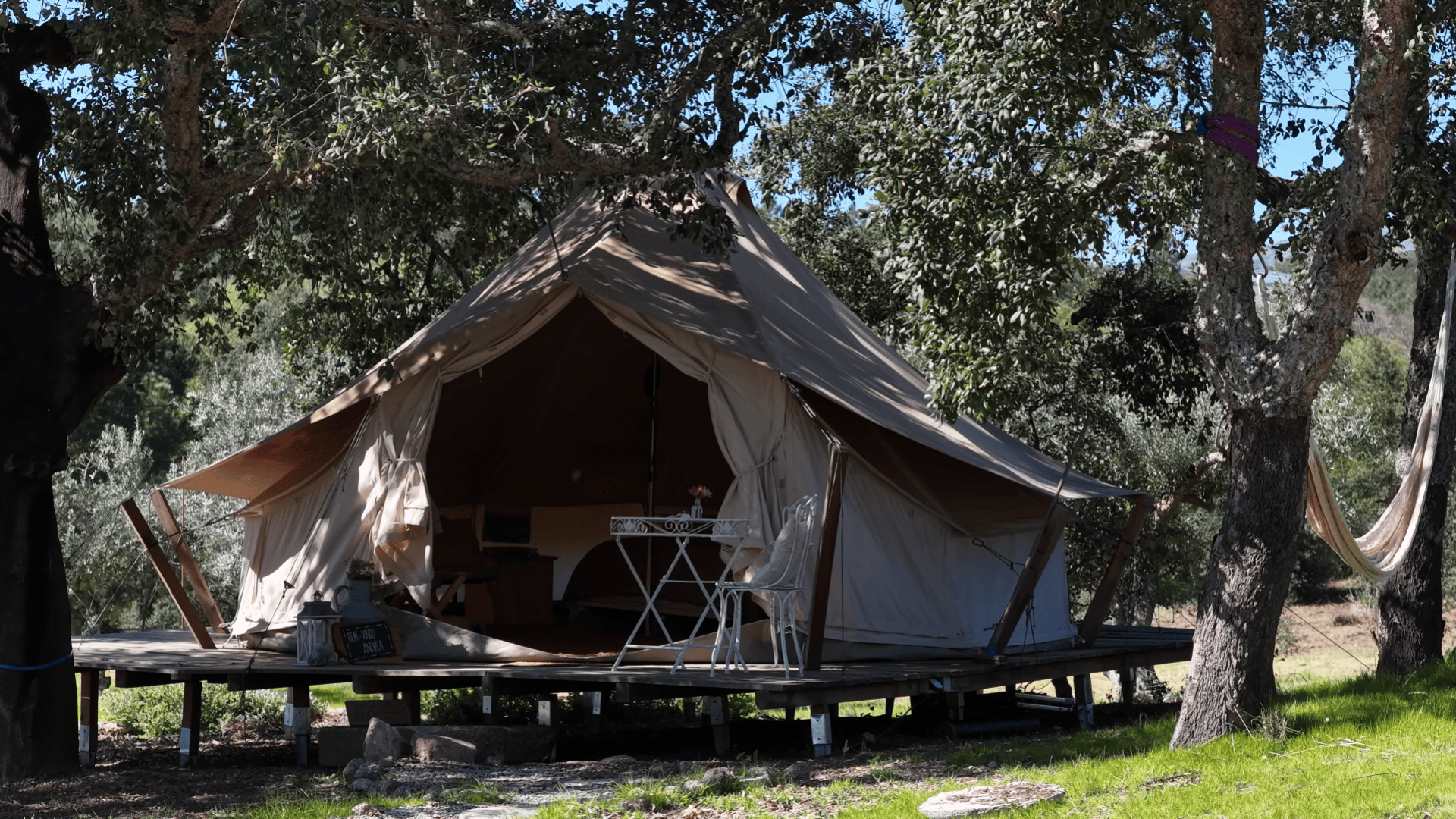 Ein großes Zelt aus Segeltuch auf einer Holzplattform unter Bäumen, davor stehen ein weißer Metalltisch und zwei Stühle. Das Zelt ist offen und gibt den Blick auf ein gemütliches Inneres mit einem Bett und kleinen Möbeln frei. Sonnenlicht fällt durch die Blätter.