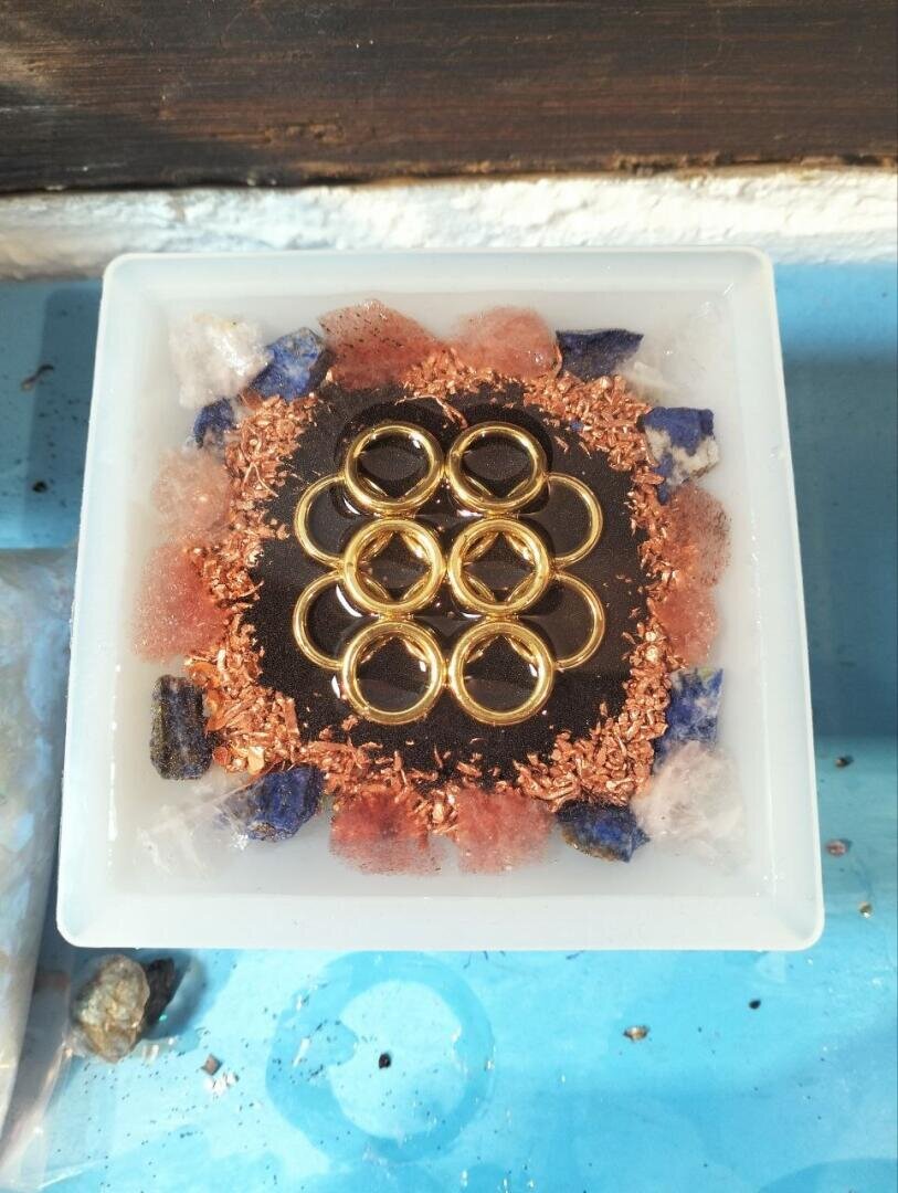 Eine quadratische Schale mit goldenen Ringen, die auf dunklem Untergrund zu einem Blumenmuster angeordnet sind, umgeben von blauen, rosa und weißen Kristallstücken sowie Kupferspänen, auf einem blauen Untergrund.
