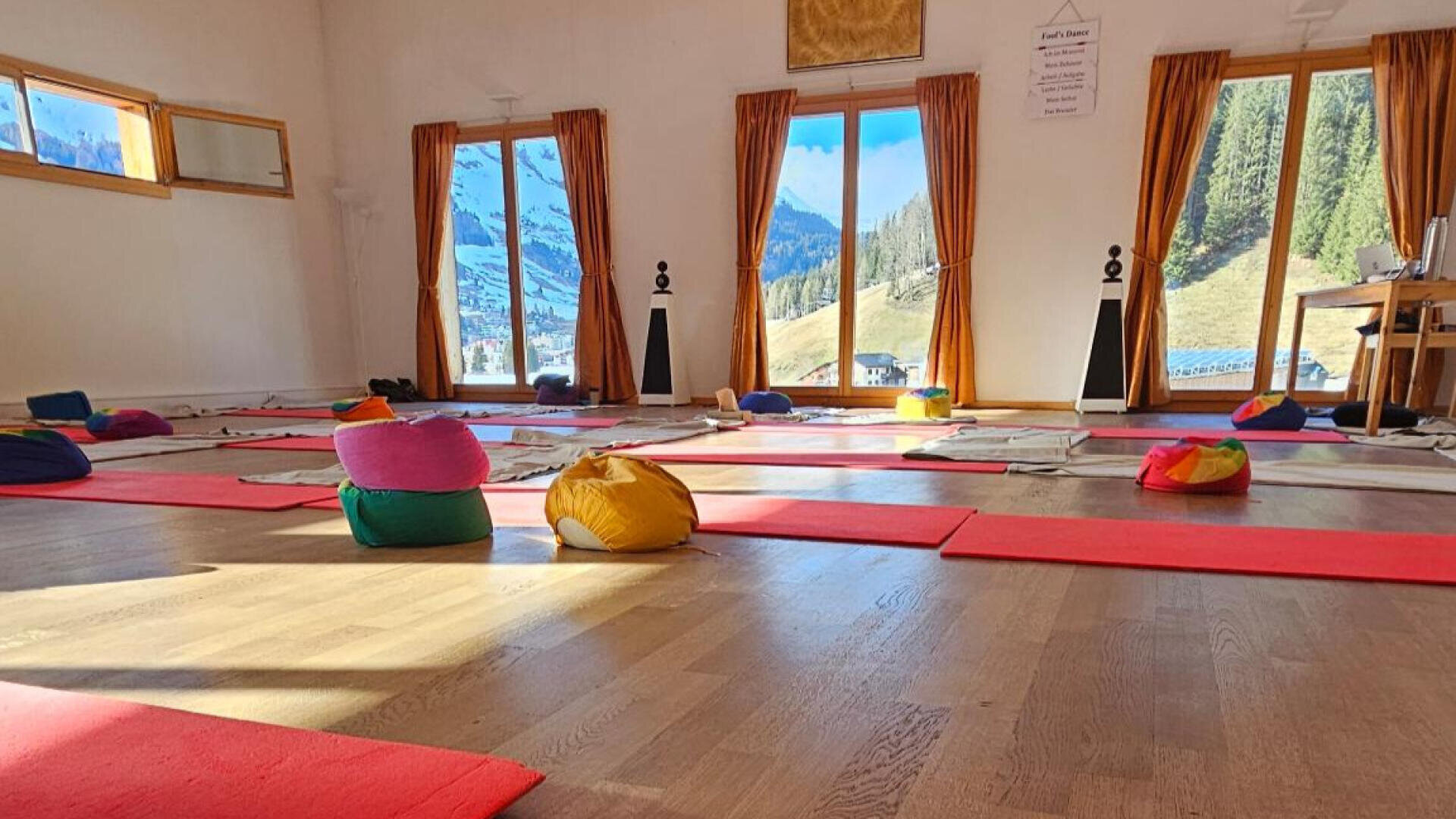 Ein helles Yogastudio mit roten Matten und bunten Kissen auf einem Holzboden. Große Fenster mit orangefarbenen Vorhängen geben den Blick auf eine malerische Berglandschaft mit Bäumen und Schnee frei.