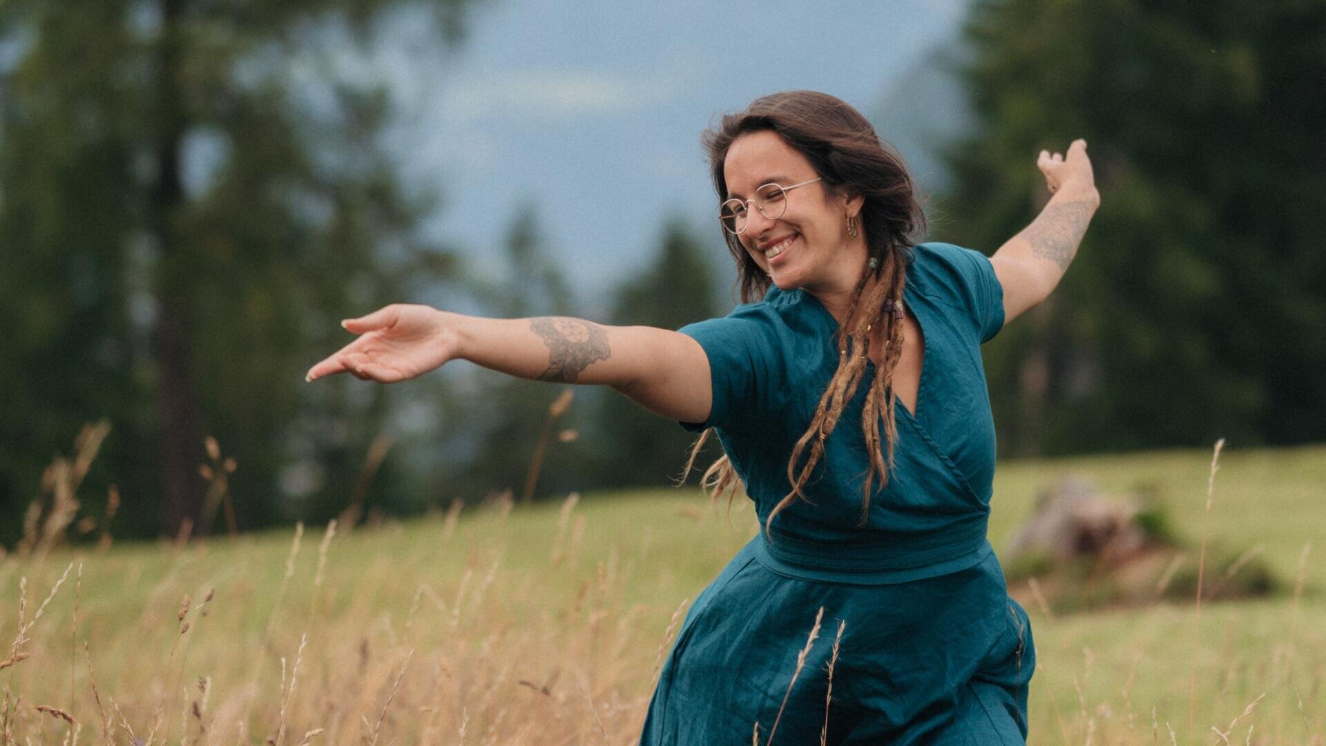 Eine lächelnde Frau mit langen Haaren und Tätowierungen, die eine Brille und ein blaugrünes Kleid trägt, steht mit ausgestreckten Armen auf einer Wiese. Im Hintergrund sind Bäume und ein bewölkter Himmel zu sehen.