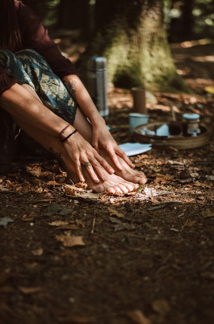 Eine Person sitzt barfuß auf dem Waldboden und berührt ihre Zehen. Daneben steht ein Tablett mit Tassen und einer Thermoskanne, während Sonnenlicht durch die Bäume fällt.