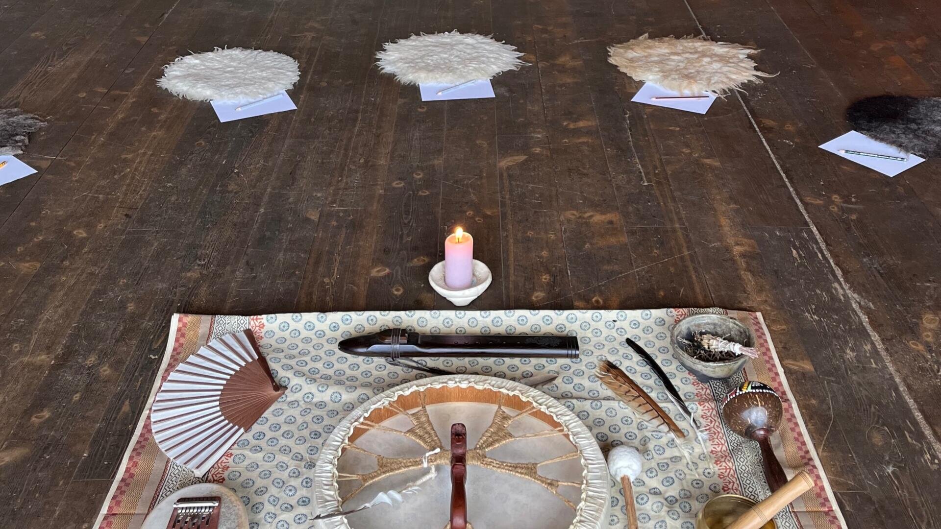 Ein ritueller Aufbau auf einem Holzboden zeigt eine brennende Kerze, eine Trommel, Federn, Muscheln und andere zeremonielle Gegenstände auf einem gemusterten Tuch. Im Hintergrund ruhen flauschige runde Objekte auf kleinen Karten.