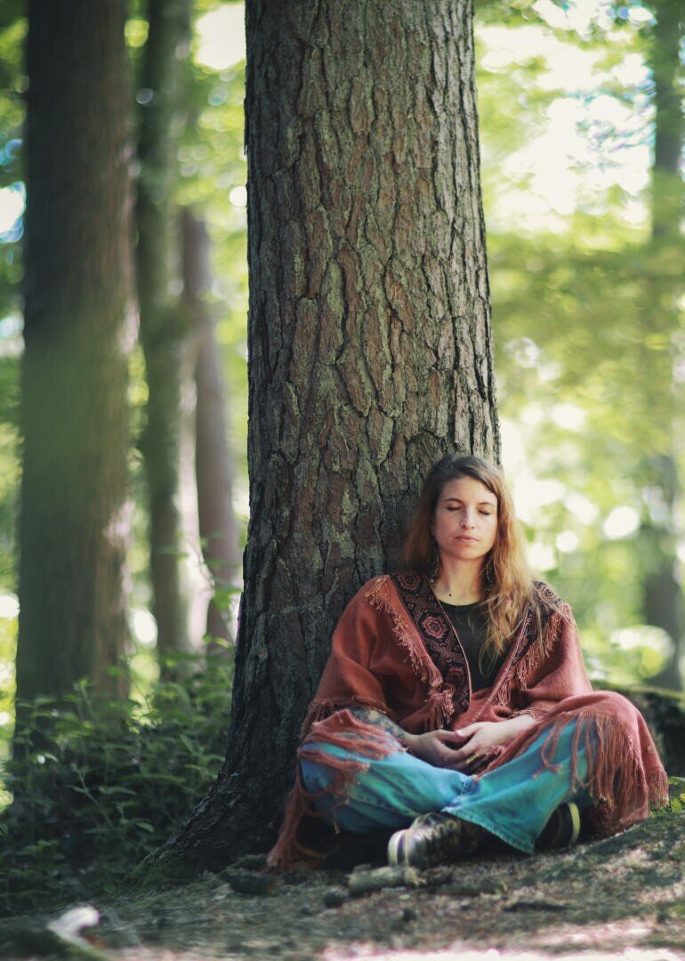 Eine Frau mit langen Haaren sitzt im Schneidersitz an einem großen Baumstamm in einem sonnenbeschienenen Wald und meditiert. Sie trägt einen rostfarbenen Schal mit Fransen und eine blaugrüne Hose mit weitem Bein, die Augen friedlich geschlossen.