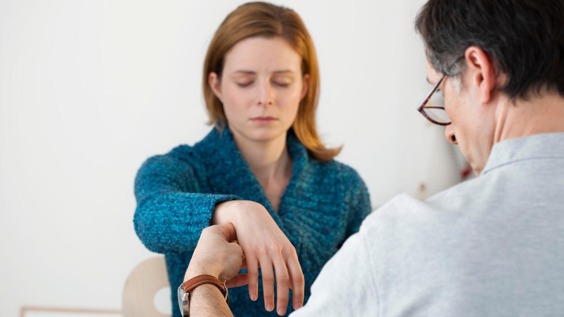 Eine Frau mit geschlossenen Augen sitzt da, während ein Mann sanft ihren erhobenen Arm hält – möglicherweise während einer Hypnotherapie- oder Entspannungssitzung – in einem hellen Raum.
