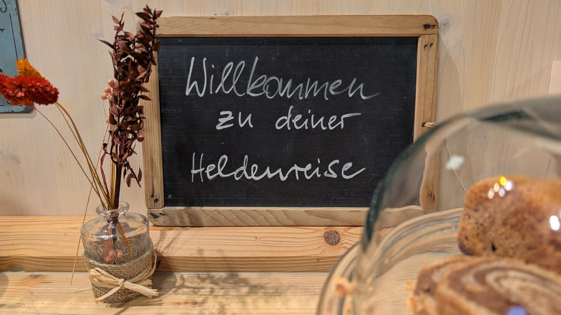 Auf einer kleinen Kreidetafel auf einem Holztisch steht "Willkommen zu deiner Heldenreise". Daneben steht eine Glasvase mit getrockneten Blumen, und im Vordergrund ist eine Torte unter einer Glaskuppel teilweise sichtbar.
