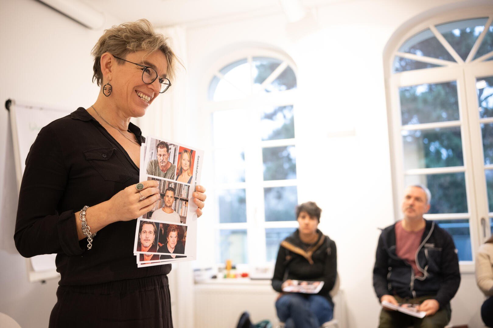 Eine lächelnde Person mit Brille hält in einem hellen Raum gedruckte Fotos hoch und spricht zu einer kleinen Gruppe von sitzenden Zuhörern.