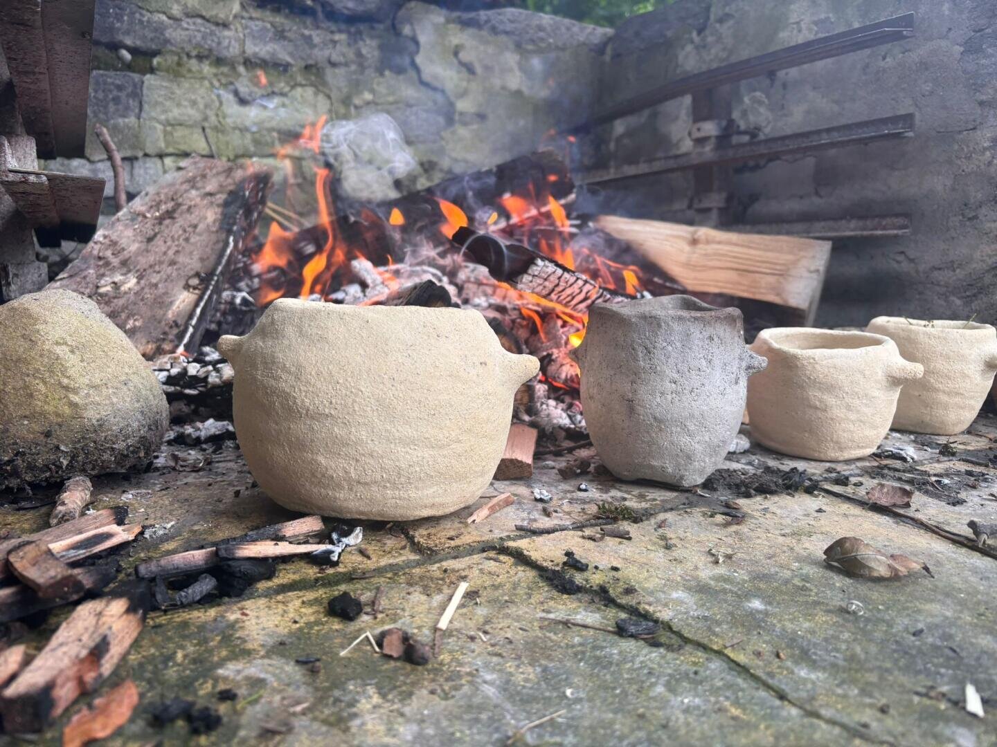 Kleine Tontöpfe stehen vor einem Holzfeuer im Freien, mit Flammen und Glut dahinter und rauen Steinmauern, die den Bereich umgeben.