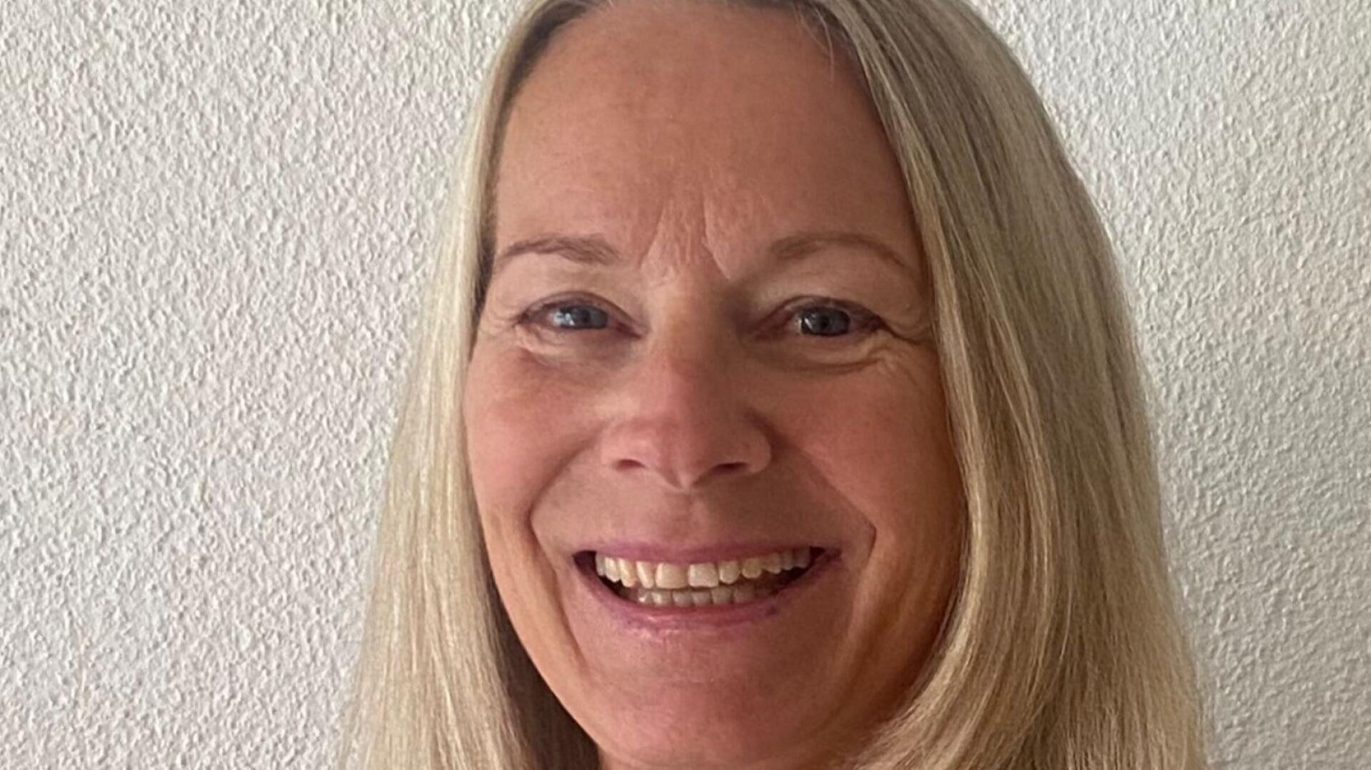 Eine Frau mit glattem, schulterlangem blondem Haar lächelt vor einer schlichten, strukturierten weißen Wand.