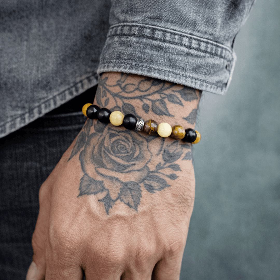 Eine Nahaufnahme einer tätowierten Hand, an der ein Perlenarmband sitzt. Das Armband besteht aus abwechselnd schwarzen, gelben und metallfarbenen Perlen. Auf der Hand ist ein großes Blumentattoo zu sehen, und die Person trägt ein graues Jeanshemd.