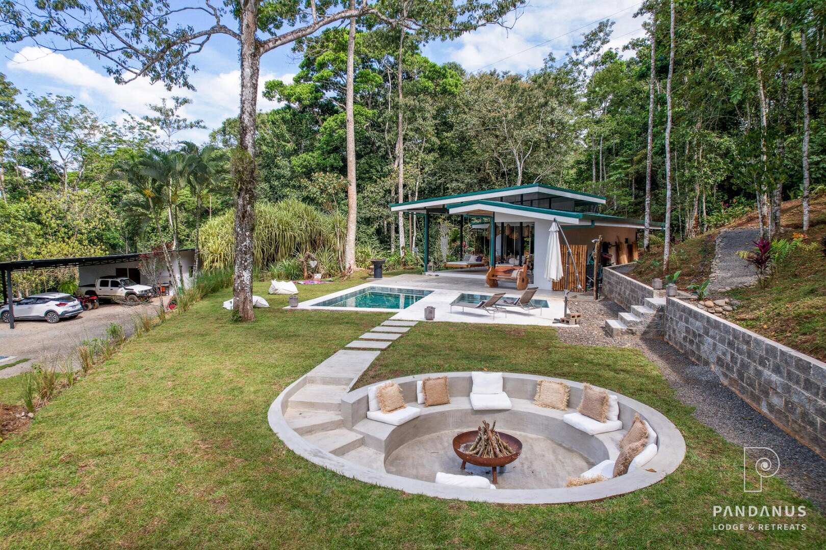 Die Pandanus Lodge & Retreats ist eine moderne Lodge mit einem Pool, einem Lounge-Bereich im Freien und einer runden, versenkten Feuerstelle mit Kissen, umgeben von üppigem Grün und hohen Bäumen.