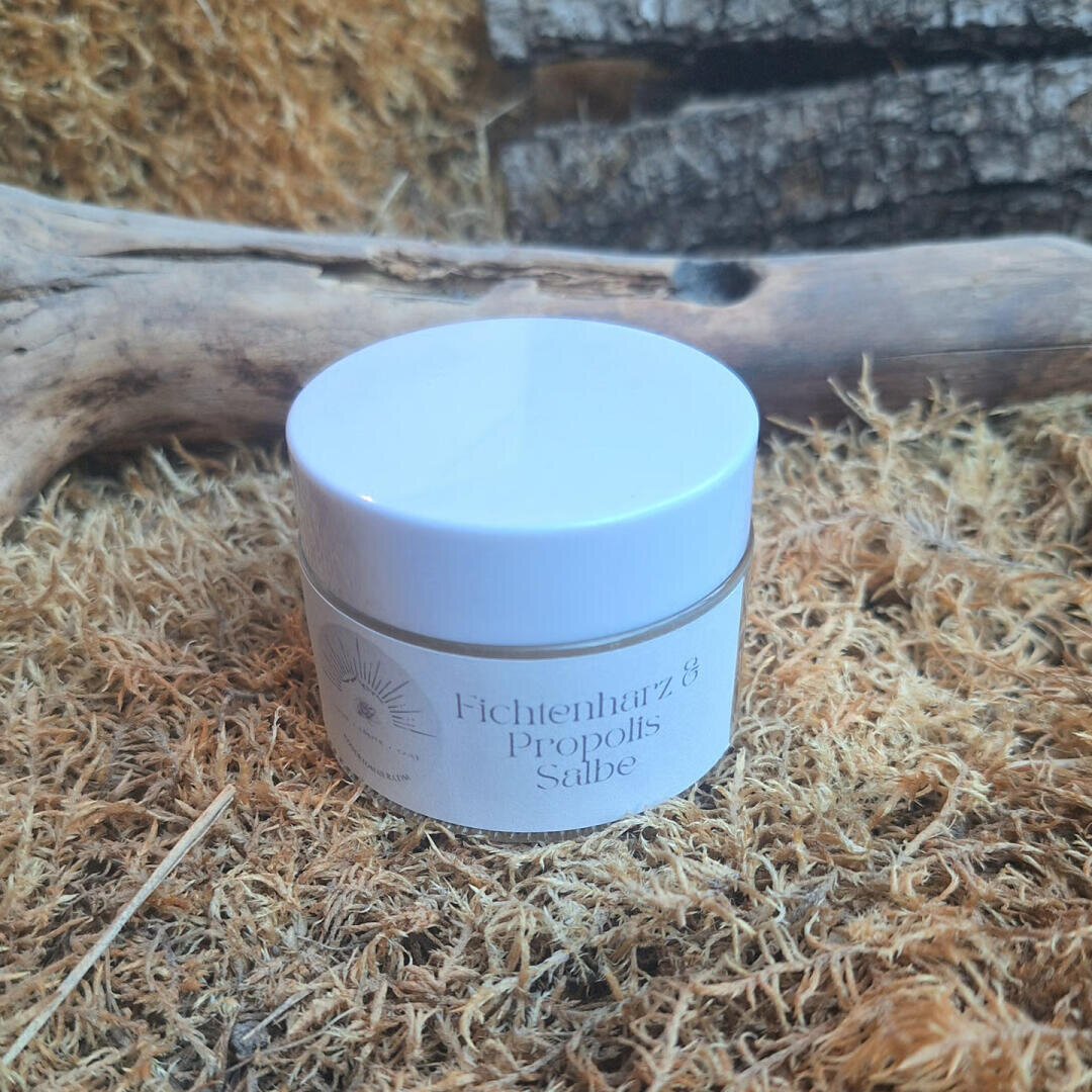 Ein kleines Töpfchen mit der Aufschrift „Fichtenharz & Propolis-Salbe“ steht auf getrocknetem Moos, im Hintergrund ist Holz zu sehen.