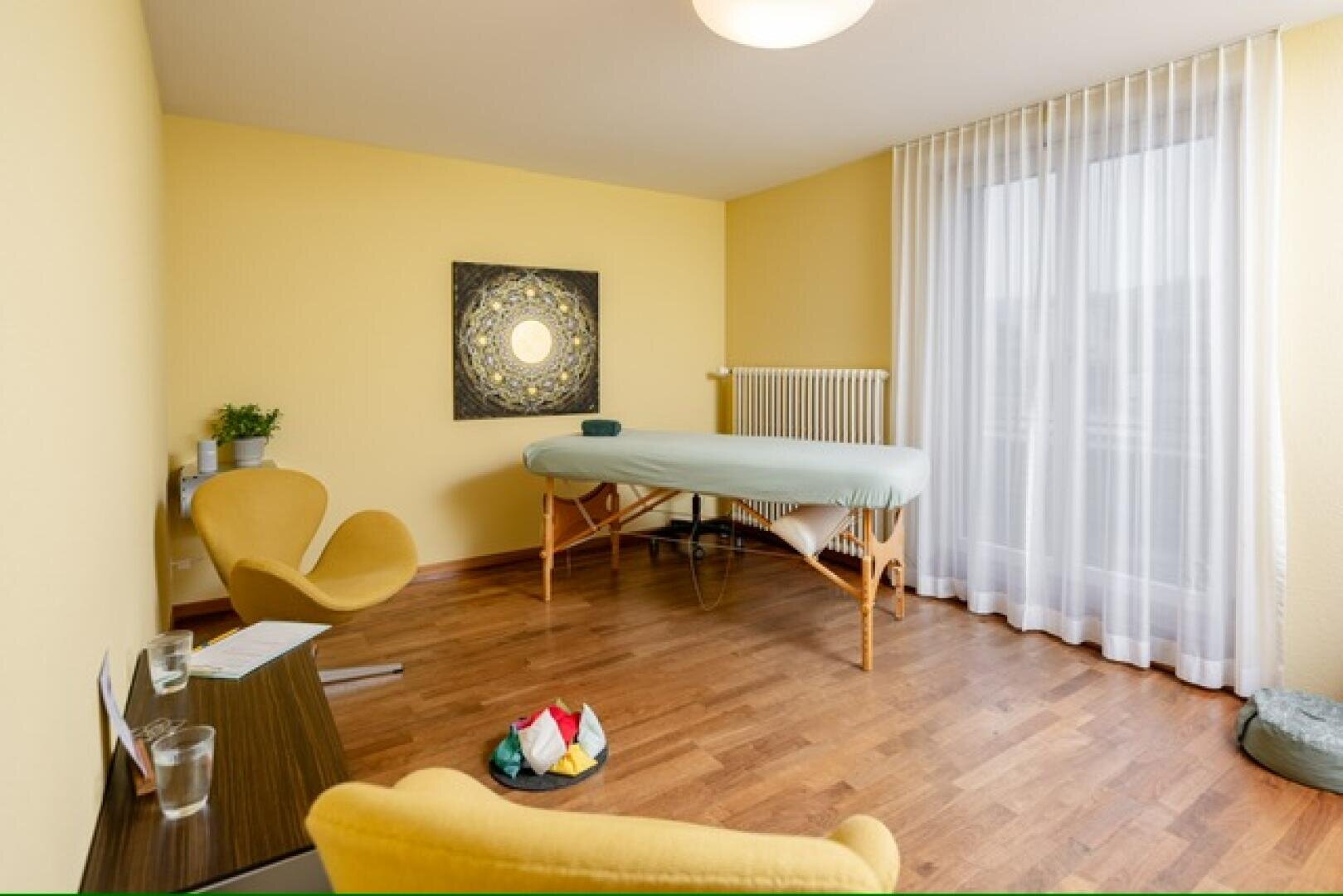 Ein heller Massageraum mit gelben Wänden, Parkettboden, einer gepolsterten Behandlungsliege, gelben Stühlen, einem Schreibtisch, transparenten Vorhängen und Mandala-Wandbildern.